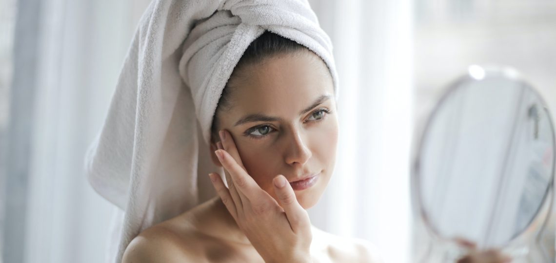 Une femme avec une serviette autour de la tête se regarde dans un miroir grossissant et semble inspecter sa peau ou appliquer un soin.